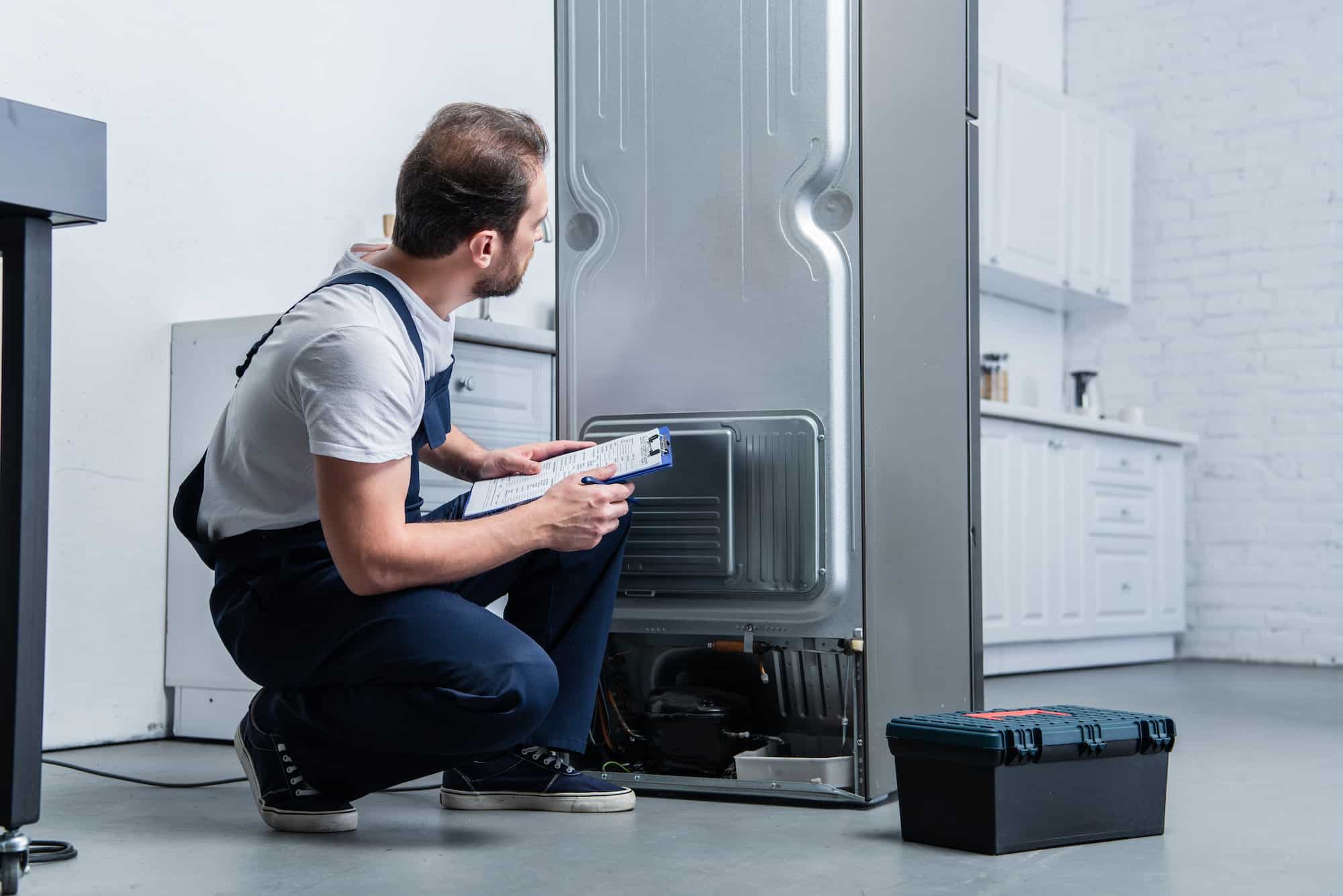 handyman-in-working-overall-writing-in-clipboard-near-broken-refrigerator-in-kitchen.jpg
